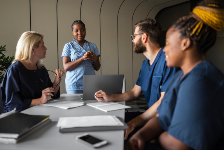 Group of healthcare professionals in a meeting room, with a nurse standing and explaining something to colleagues, while two others sit at the table with laptops and notebooks.